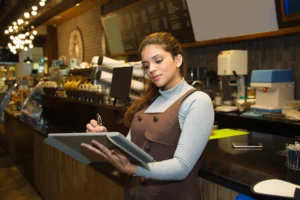 A young woman in a brown uniform and grey turtleneck stands behind a cafe counter while writing in a large ledger. The background features a warm, modern coffee shop interior with industrial lighting and professional espresso equipment.
