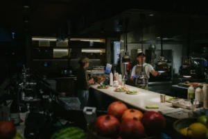 This image depicts a bustling restaurant kitchen where staff are busy plating fresh salads under warm heat lamps. In the foreground, vibrant pomegranates and other fruits add a splash of color to the moody, low-lit atmosphere.