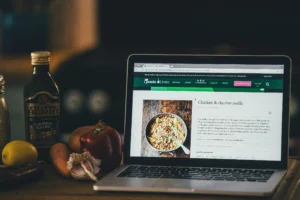 A laptop open to a Jamie Oliver recipe for "Chicken & chorizo paella" sits on a kitchen counter next to fresh ingredients. The surrounding ingredients include a bottle of olive oil, a lemon, a carrot, a red bell pepper, and a head of garlic.
