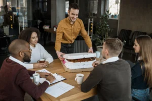 A smiling man in a yellow shirt places several pizza boxes on a wooden conference table for four colleagues during an office meeting. The diverse group of professionals sits around the table with coffee mugs and business documents, looking on as the food is delivered.