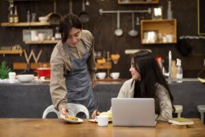A man wearing a grey apron serves a small pastry to a woman working on her laptop at a wooden table. They are in a cozy, rustic cafe or kitchen setting with dark wood shelves and cooking utensils in the background.
