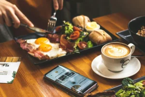 A person uses a knife and fork to eat a breakfast plate featuring fried eggs, bacon, and a fresh side salad. A latte with heart-shaped foam sits nearby on the wooden table next to a smartphone and a travel brochure.