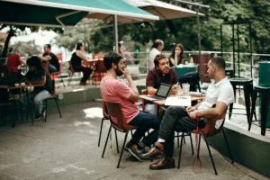 Three men sit around a table at an outdoor patio cafe, deeply engaged in a conversation with a laptop and notebooks open before them. In the background, other patrons sit at tables under green umbrellas, creating a lively and relaxed atmosphere.