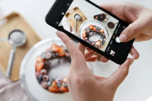 A person holds a smartphone to capture a top-down photograph of a round chocolate cake decorated with fresh berries and powdered sugar. The phone screen displays the framed shot of the cake alongside baking tools and ingredients arranged on a white surface.