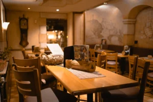 Warm, ambient lighting fills this rustic dining room, which features wooden tables and chairs set against walls adorned with faded murals. A long table in the foreground holds menus and a decorative woven basket, creating an inviting, cozy atmosphere.
