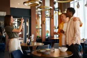 In the file, a woman is capturing a photo of two men posing with wine glasses in a modern restaurant. The setting features elegant gold ring light fixtures and circular tables, creating a warm and social atmosphere.