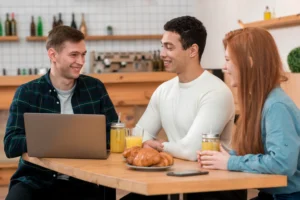 Three friends sit together at a wooden table in a cafe, smiling and interacting around a laptop, pastries, and drinks. A man in a plaid shirt looks at the laptop on the left, while a man in a white sweater and a woman with red hair converse beside him.