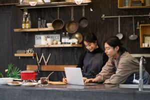 Two people wearing aprons stand behind a kitchen counter while looking at a laptop screen together. One person leans over the computer while the other takes notes with a pen in a rustic, modern cafe setting.