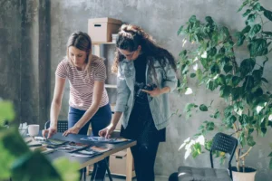 Two women collaborate in a sunlit, rustic studio, reviewing printed photographs spread across a dark table. One woman in a striped shirt leans over the table to arrange the prints, while the other, holding a camera, looks down to assist her.