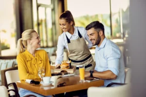 A smiling waitress wearing an apron serves a burger and fries on a wooden tray to a happy couple sitting at an outdoor restaurant table. The couple, a woman in a yellow blouse and a man in a light blue shirt, look up at the waitress and smile warmly.