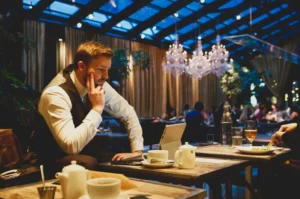 A man in a waistcoat sits at a table in a dimly lit, upscale restaurant, appearing deep in thought while looking at a tablet. The background features elegant chandeliers and a glass ceiling that reveals a dark, evening sky.