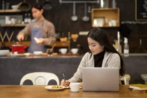 In the image, a young woman sits at a wooden table in a cafe, focused on writing in a notebook next to her open laptop. In the blurred background, a man in an apron works behind the counter of the rustic, industrial-style kitchen.