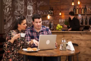 A man and a woman sit at a round wooden table in a rustic cafe, focused on a laptop screen while enjoying coffee and croissants. The background features a warmly lit bar area with a barista, complemented by chalkboard wall art and hanging Edison bulbs.