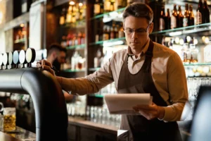A focused man wearing an apron and glasses stands behind a bar counter while reviewing a paper inventory list. The background is filled with rows of glass bottles on illuminated shelves, creating a warm and busy atmosphere.