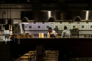 Two chefs in blue aprons and caps work behind a dimly lit restaurant counter under three low-hanging dome lights. The background features a tiled wall and stacks of ceramic plates, creating a focused, industrial kitchen atmosphere.