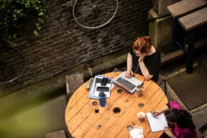 An overhead shot shows two women working at a large, rustic wooden cable spool table in an outdoor courtyard. One woman typing on a laptop and another writing in a notebook are surrounded by brick walls decorated with bicycle wheels and ivy.