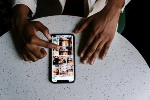 An overhead shot shows a person with a dark complexion using their left hand to scroll through a photo grid on a smartphone. The phone rests on a speckled, light-colored round table next to their resting right hand.