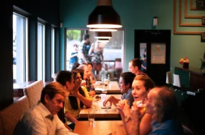 A group of people are gathered around long wooden tables in a dimly lit, modern restaurant or taproom. Warm pendant lights hang overhead, casting a glow on the patrons as they engage in lively conversation over drinks.
