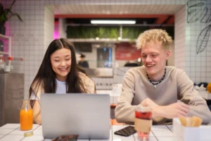 A young woman with long dark hair and a young man with blonde, curly hair sit side-by-side at a tiled table, smiling while looking at a laptop screen. They are inside a modern cafe or restaurant, with drinks and a smartphone placed on the table in front of them.
