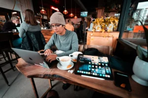 A person wearing a beanie and glasses sits at a live-edge wooden table in a coffee shop, focused on typing on a laptop. Their workspace is filled with tech gear, including a large tablet, a smartphone, and wireless earbuds, alongside two white coffee cups.