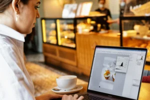 A woman sits at a table in a cafe, focusing on her MacBook Pro while editing a photo of pastries in Adobe Photoshop. Next to her laptop, a hot cup of coffee sits on a saucer, with a warm-lit bakery display counter visible in the background.
