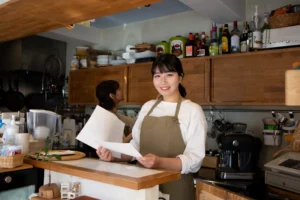 A young East Asian woman wearing a white long-sleeved shirt and an olive green apron stands behind a wooden cafe counter, smiling at the camera while holding papers. In the background, another woman works near wooden cabinets filled with bottles, plates, and an espresso machine.