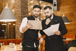 Two men with beards stand together in a dimly lit restaurant or cafe, reviewing a digital tablet and printed papers. The man on the left holds up the tablet while the man on the right listens thoughtfully, clutching documents in one hand and touching his tie with the other.