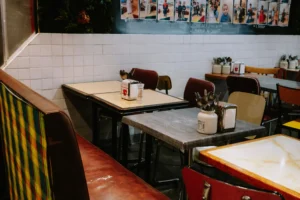 An indoor shot of a retro-style diner or café features small square tables, mismatched chairs, and a red vinyl booth with a colorful plaid backrest. A white tiled wall lines the background, topped by a chalkboard displaying menu items and a row of photographs.