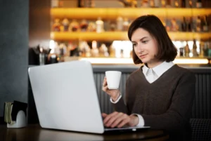 In the image, a woman with short brown hair sits at a table while typing on a laptop and holding a small white coffee cup. She is dressed in a brown sweater over a collared white shirt, with a warmly lit bar area visible in the softly blurred background.