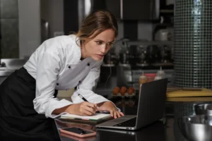 A female chef wearing a white double-breasted uniform and black apron leans over a stainless steel kitchen counter. She focuses intently on a laptop screen while writing notes in a spiral notebook, with a smartphone, mixing bowls, and a carton of eggs nearby.