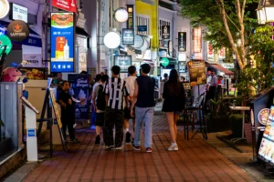 A group of people walks away from the camera down a lively, brick-paved pedestrian street lined with illuminated restaurant and bar signs at night. The bustling urban scene features a mix of diverse storefronts, glowing advertisements, and outdoor dining areas under a canopy of trees.