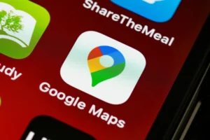The image shows a close-up macro shot of a smartphone screen displaying the Google Maps application icon. The white, rounded-square icon features the colorful Google pin logo and is situated on a dark red background alongside parts of neighboring app icons.