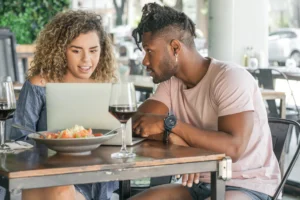 The image shows a young couple sitting at an outdoor restaurant table while looking intently at a laptop screen. In front of them, a fresh salad and two glasses of red wine are served on the wooden table.