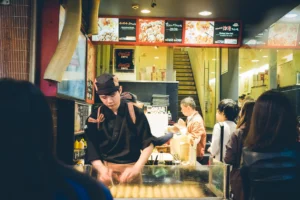 In the image, a young chef in a black uniform and head covering is focused on preparing takoyaki on a large griddle at an open-front street food stall. Customers and passersby stand outside the brightly lit shop, which features vibrant menu boards and a staircase leading upstairs.