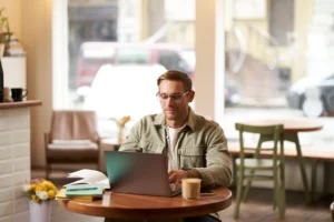 A young man with glasses and a green button-down shirt sits at a wooden cafe table, working on a laptop. An iced coffee, open notebook, and pens are placed on the table next to him against a blurred background of the coffee shop interior.