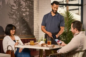 A man at a restaurant table uses his smartphone to make a contactless payment on a handheld terminal held by a server. A woman sits across from him, holding a glass of wine and watching the interaction.
