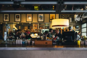 The image shows a bustling indoor scene from behind a bar counter, looking toward patrons and staff in a dimly lit restaurant or brewery. A warm, large drum pendant light illuminates customers conversing near the counter, while framed posters hang on the wall in the background.