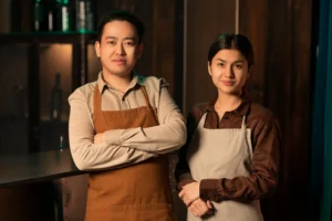 A medium shot of a man and a woman standing side-by-side behind a bar counter, wearing aprons and button-down shirts while posing for the camera. The background features a dimly lit, rustic wooden interior with blurred bottles on a shelf.