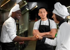 Three kitchen staff members are smiling and interacting in a commercial kitchen setting. A Black chef on the left and a female chef on the right, both wearing white chef uniforms and hats, are doing a fist bump while a man in a black apron watches with his arms crossed.