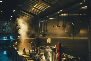A dimly lit professional kitchen features a large plume of steam rising from a cooking station on the counter. Various stainless steel pots, pans, and hanging utensils are illuminated by warm overhead lights against a metallic wall.