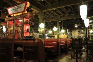 An indoor view of a vintage-style diner featuring wooden booths with red leather seating under warm, hanging light fixtures. A retro marquee sign advertises strawberry banana pudding above a souvenir display case filled with bottled sauces and merchandise.