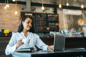 In the image, a woman in a white blouse sits at a table inside a warmly lit cafe, looking at her laptop screen. She gently smiles while holding a white coffee cup, with a chalkboard menu and industrial hanging lights visible in the blurred background.