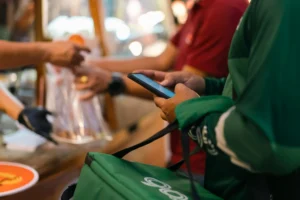 A delivery courier wearing a green jacket holds a smartphone while waiting at a food stall. In the blurred background, a merchant hands over a packaged order.