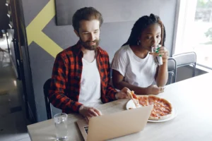 In the image, a man with a beard wearing a red flannel shirt looks at a laptop while holding a slice of pepperoni pizza. Next to him, a Black woman with long braids drinks a glass of water while looking down at the screen.