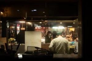 In the image, a worker wearing a green cap is seen from behind through a window as they work inside a dimly lit shop or restaurant kitchen at night. Out-of-focus customers and the bright lights of a bustling street or market are visible through the glass in the background.