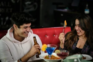 A smiling young man in a white hoodie holds a beer bottle while looking at a woman sitting next to him on a red booth. The woman laughs as she holds up a fork with food, with colorful cocktails and dishes spread across the table in front of them.
