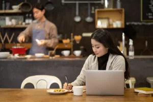 A young woman with long dark hair sits at a wooden table in a cafe, focusing on a laptop while writing in a notebook with her right hand. In the blurred background, a man wearing an apron works behind the kitchen counter near shelves and cooking utensils.