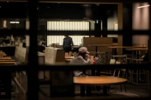 A person wearing a cap sits at a wooden table inside a dimly lit cafe, focused on an open laptop. The candid shot is framed through a dark window, with empty seating and a blurred figure visible in the background.