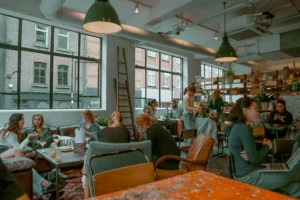 A bustling cafe with large industrial-style windows is filled with patrons sitting at tables and on leather sofas. Several large green dome pendant lights hang from the exposed ceiling, illuminating people chatting, drinking, and working on laptops.