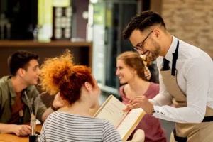 A smiling waiter wearing glasses, a white shirt, and an apron stands at a restaurant table, pointing to options on a menu for a group of friends. A woman with bright red hair looks up at him from the foreground while two other patrons chat and smile in the background.